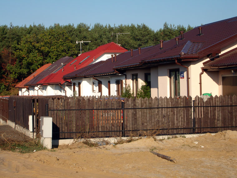 Szereg nowo wybudowanych domów jednorodzinnych z brązowymi i czerwonymi dachami, widzianych zza drewnianego ogrodzenia, na tle lasu.